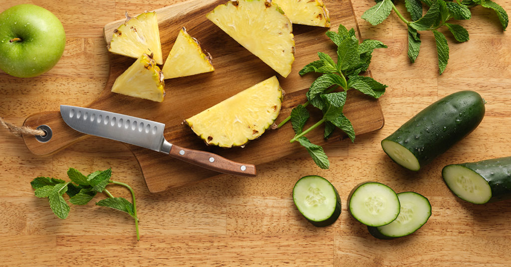 ingredients to make cold pressed green juice with mint, pineapple, green apple and cucumber on a wooden cutting board with a knife