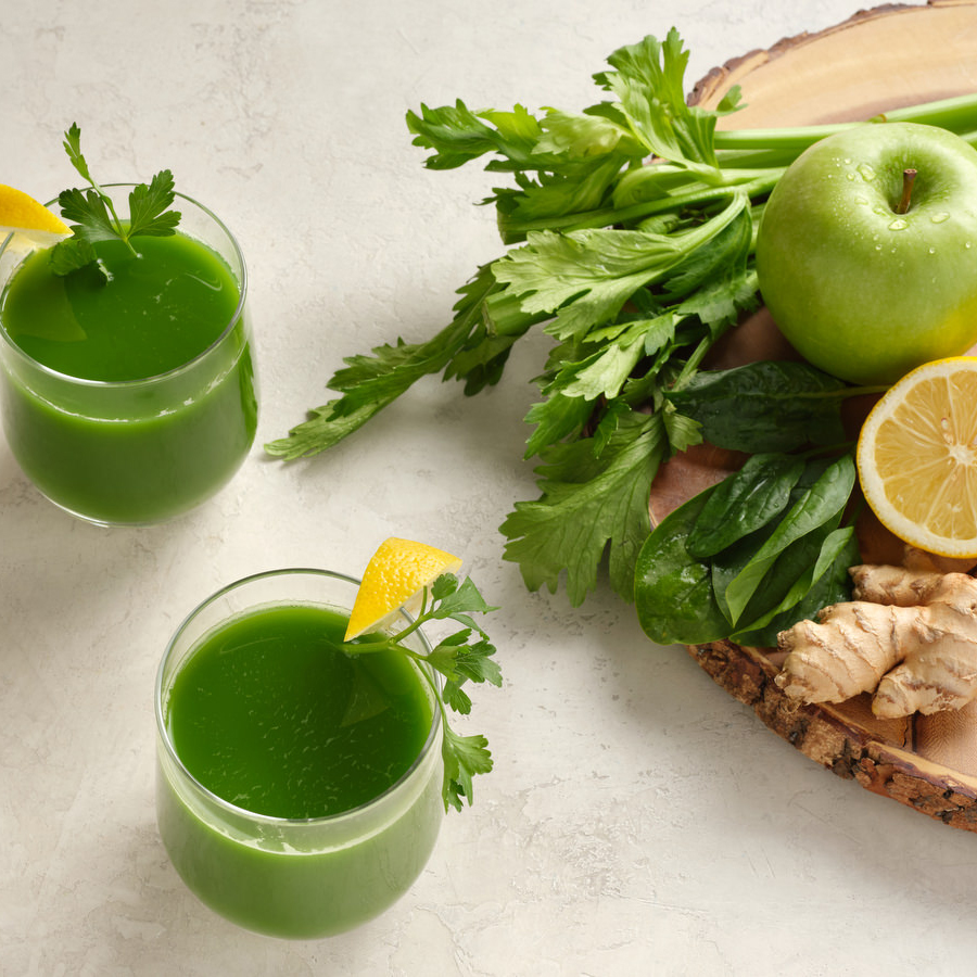 two glasses of green spinach juice on a white table surrounded by ingredients, apple, lemon, ginger, celery and parsley