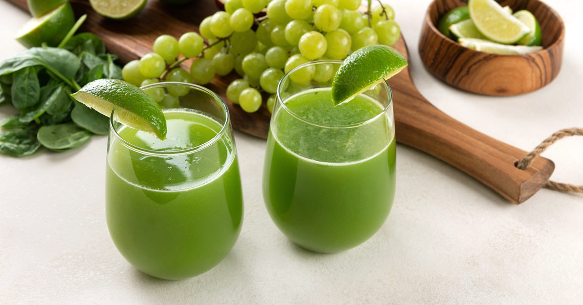 two glasses of apple limeade on a white surface surrounded by ingredients grape, spinach, lime, and apple