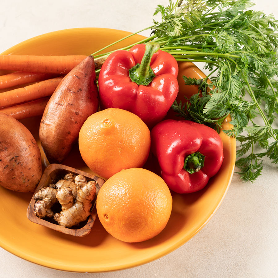 Ingredients to make red bell pepper juice including carrots, sweet potato, orange and ginger in an orange bowl